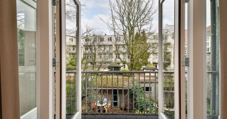 Widok z wnętrza mieszkania przez otwarty balkon, na zielony teren między blokami