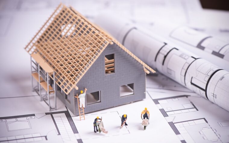 Makieta domu w trakcie budowy stojąca na arkuszach papieru z planami budowy. Obok domu figurki robotników
